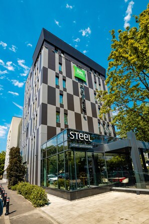 A hotel with a checkered facade, a visible sign for 'Ibis', and a glass entrance with outdoor seating.
