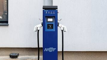 Una estación de carga para vehículo eléctrico azul, con dos cables conectados, sobre una superficie de concreto frente a una pared blanca.