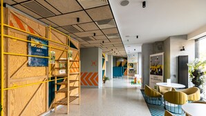 A modern interior with a central corridor, wooden paneling on one side, and a seating area with yellow chairs and a white table.