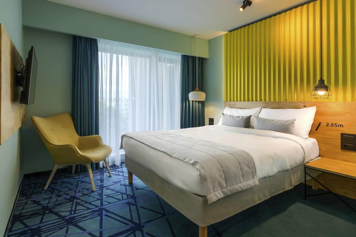 A hotel room with a bed, a chair, a desk, and a large window with curtains.