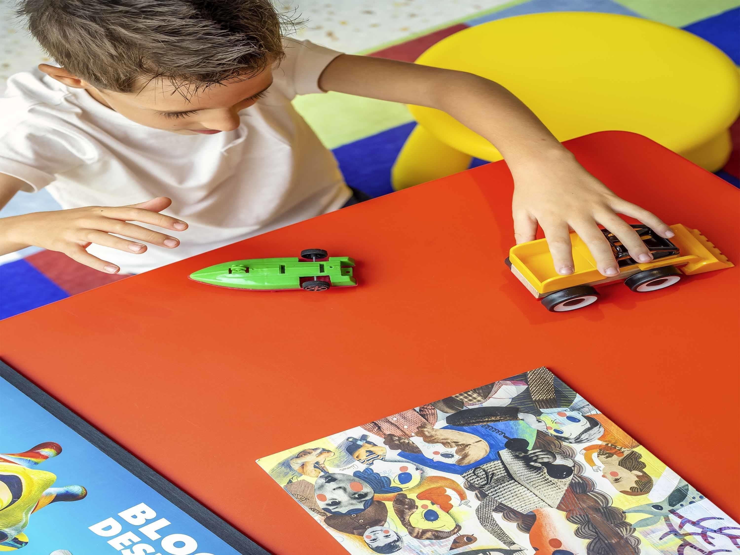 A child playing with toy vehicles on a table with a colorful play mat and a book featuring cartoon characters.
