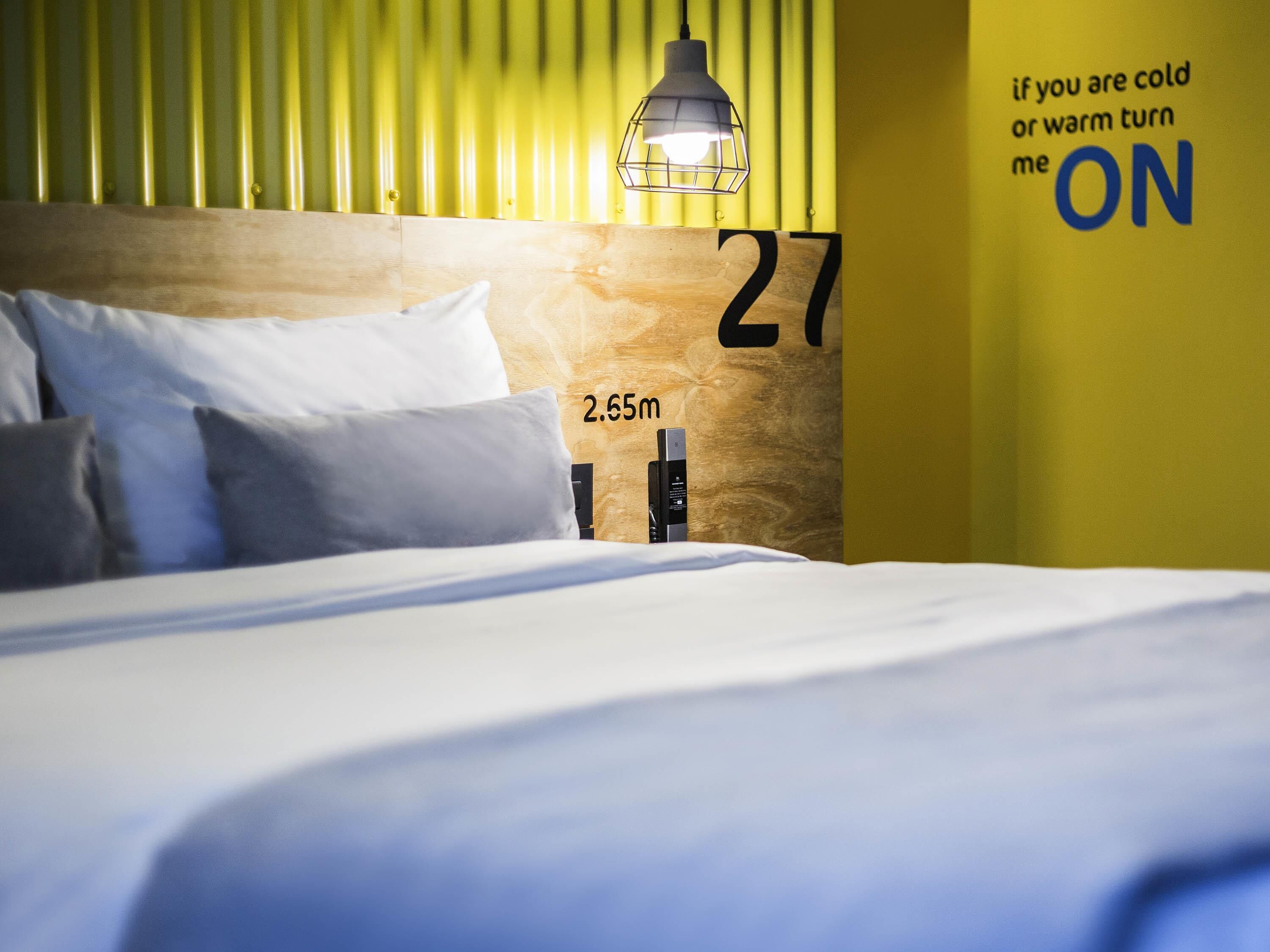 A bed with white bedding, a wooden headboard with a number, a lamp, and a wall with a temperature control sign.