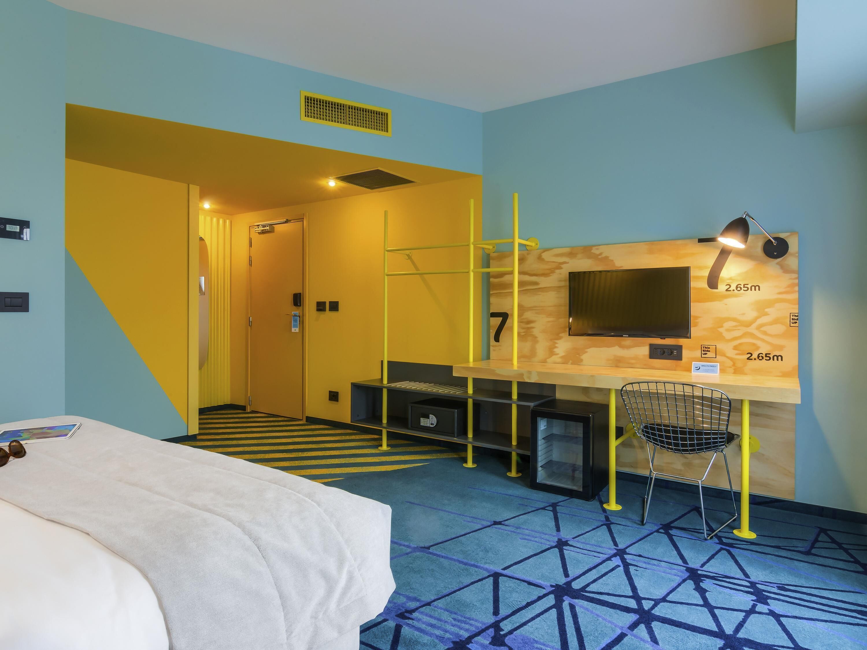 A hotel room with a bed, a desk with a chair, a TV, and a patterned carpet.