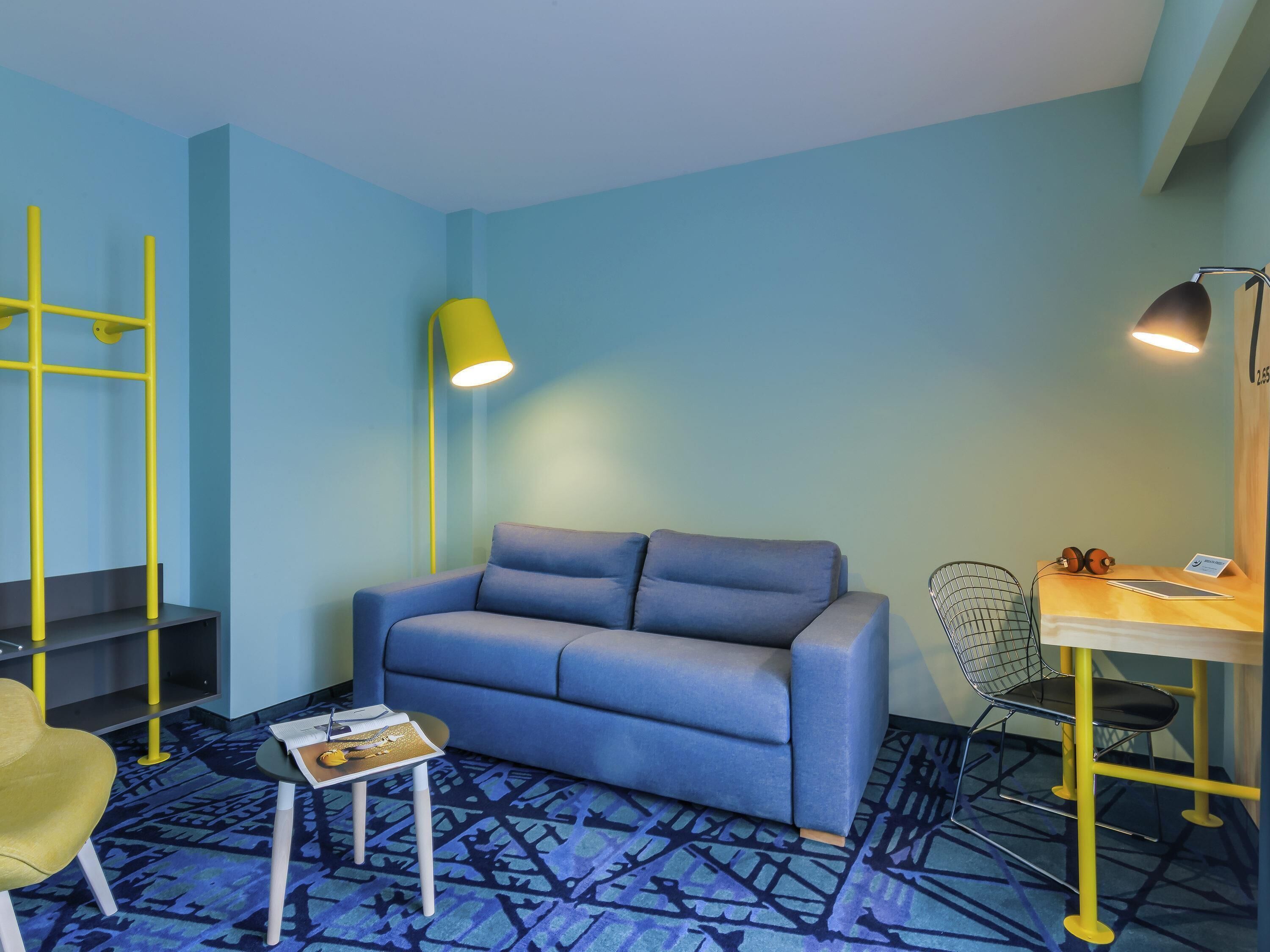 A room with a blue sofa, a small table with a magazine, a yellow floor lamp, a black chair, and a desk with a lamp and headphones.