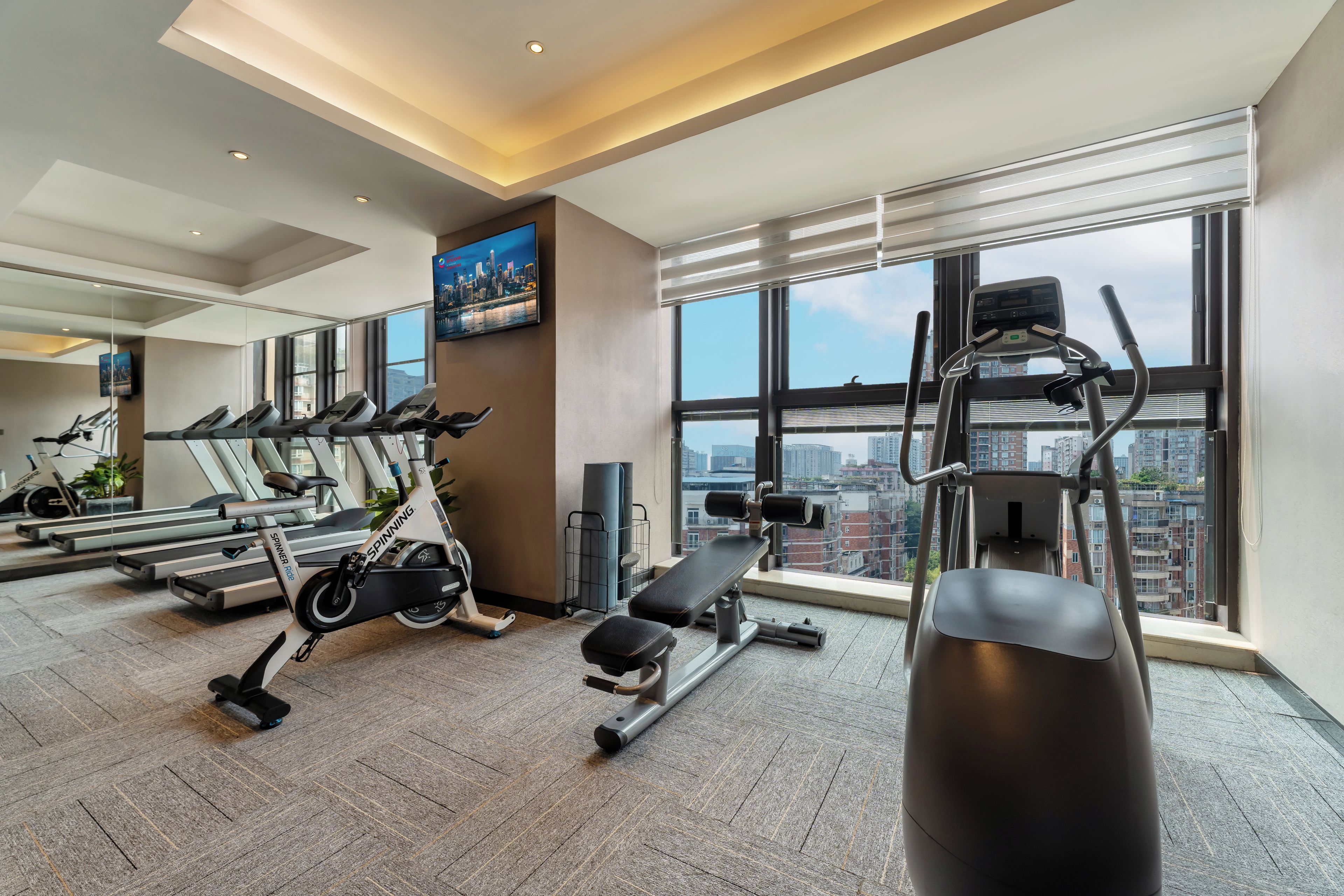 A modern gym with treadmills, a spinning bike, and weight equipment, featuring large windows and a city view.
