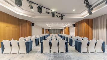 Sala de conferências com fileiras de mesas e cadeiras, com iluminação pendente e um palco.