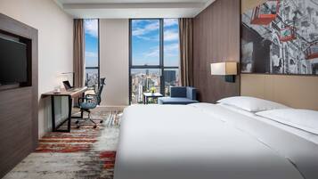 A hotel room with a large bed, a desk with a laptop, a chair, a TV, and a city view through large windows.