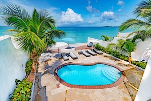 A swimming pool with lounge chairs and umbrellas, surrounded by palm trees and a view of the ocean.