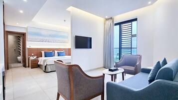 A hotel room with a bed, a TV, a sofa, and a chair. There is a window with curtains and a view of the outside.