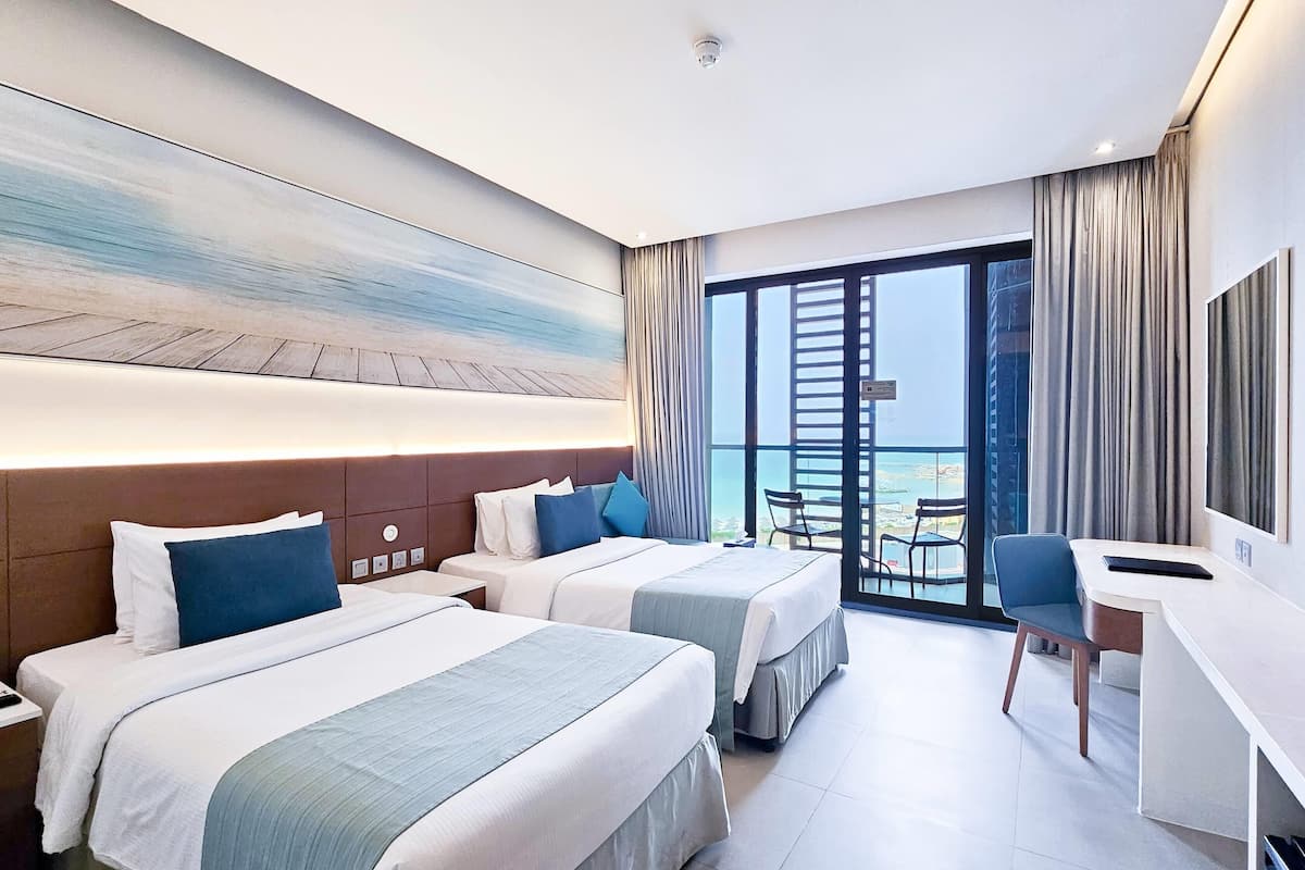 Een hotelkamer met twee bedden, een bureau en een groot raam met uitzicht op het strand.