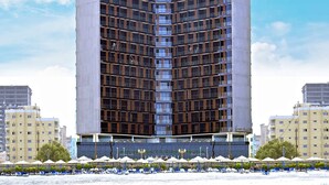 Un hotel di grandi dimensioni dal design moderno, situato vicino a una spiaggia con ombrelloni e lettini.