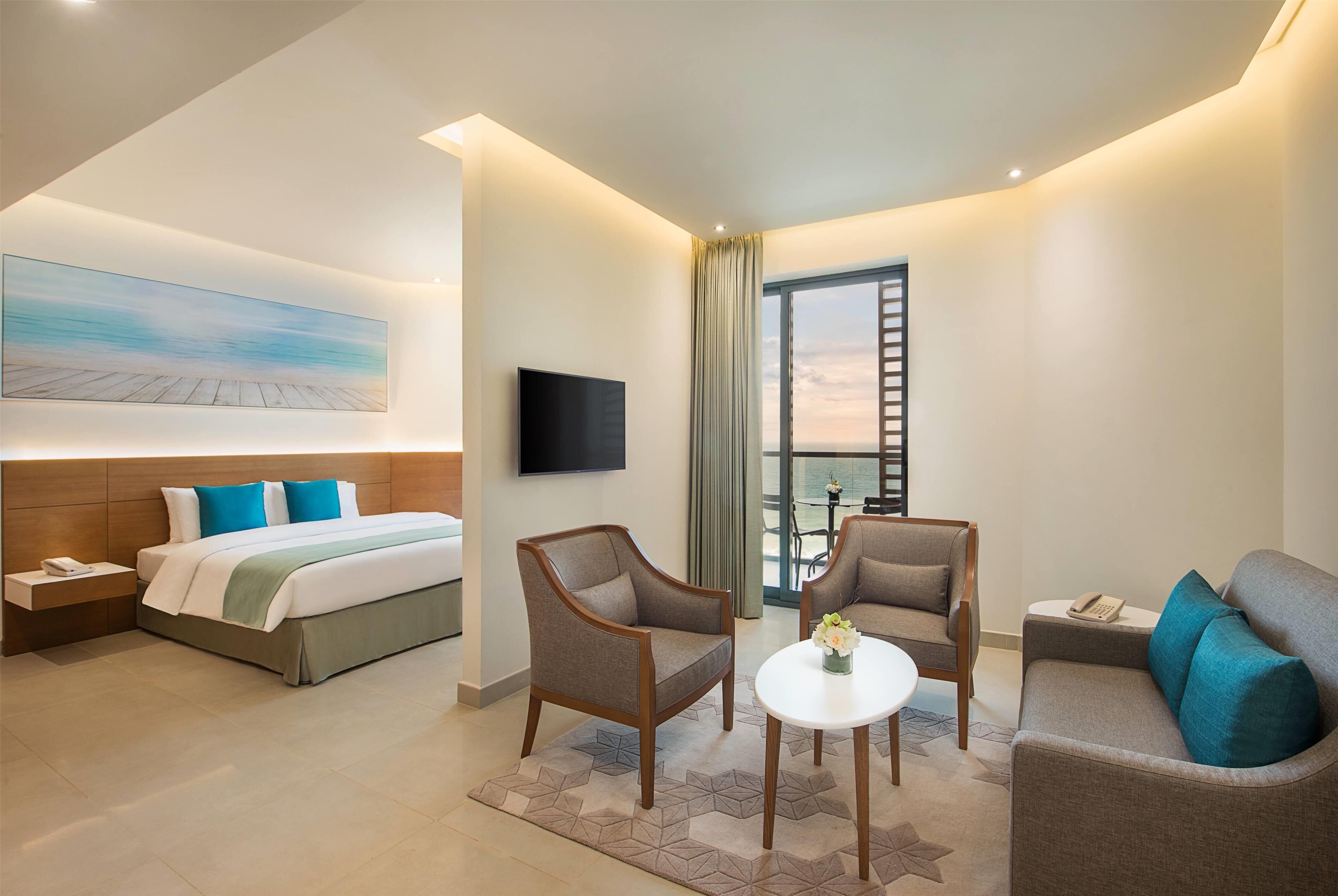 Une chambre d’hôtel moderne équipée d’un lit, de deux fauteuils, d’un canapé, d’une petite table, d’une télévision et d’un balcon avec vue.