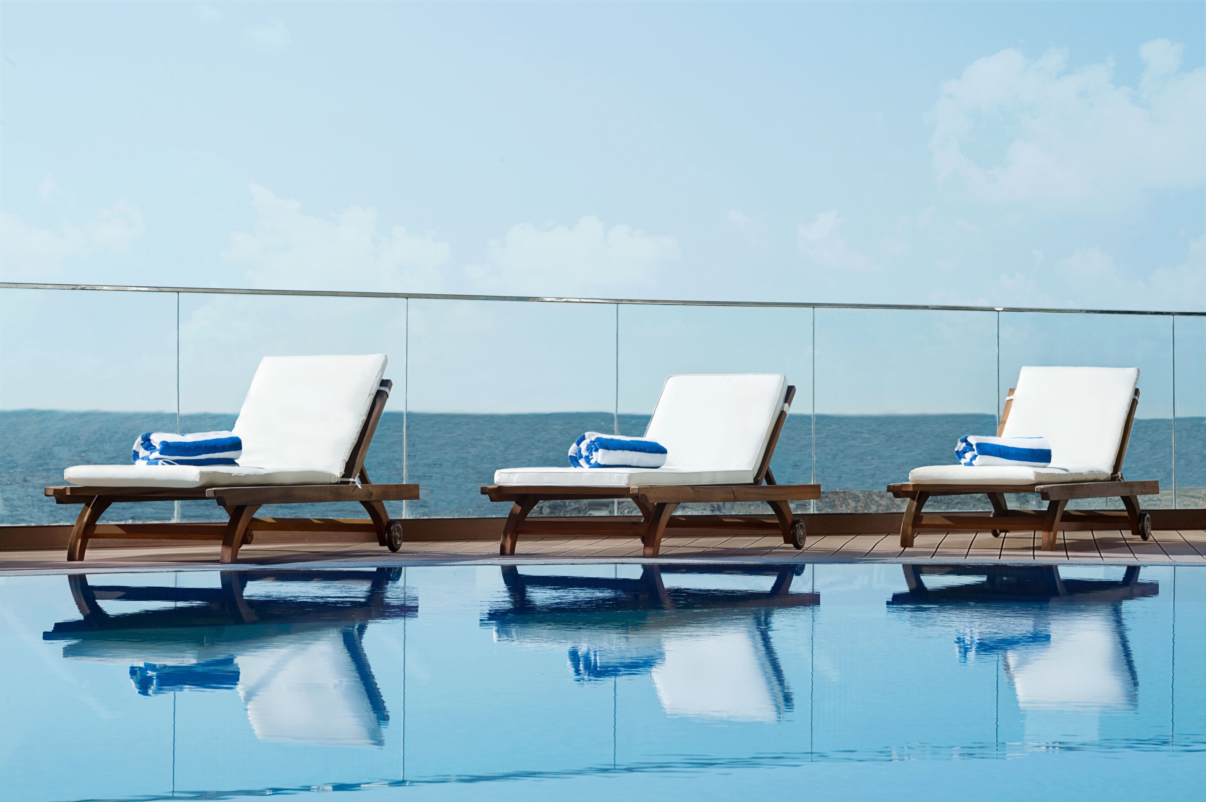A poolside area with three white lounge chairs, each with a blue towel, overlooking a body of water.
