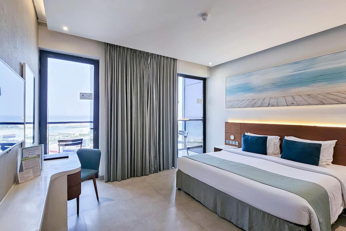 Een hotelkamer met een bed, een bureau en een stoel. Er is een groot raam met gordijnen en een schilderij aan de muur.