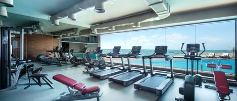 A gym with treadmills, weight machines, and dumbbells, overlooking a pool and beach.