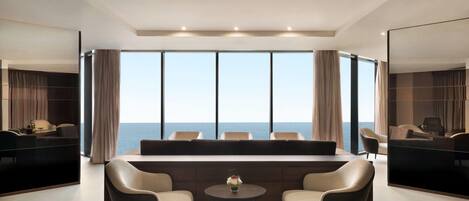 Habitación de hotel moderna con un ventanal grande que ofrece vistas al mar, un sofá, dos sillones y una mesita con un florero.