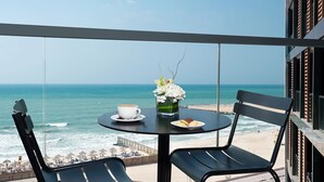 Un balcone con tavolo, sedie, una tazza di caffè, un piatto di biscotti e un vaso di fiori, vista su una spiaggia e l'oceano.