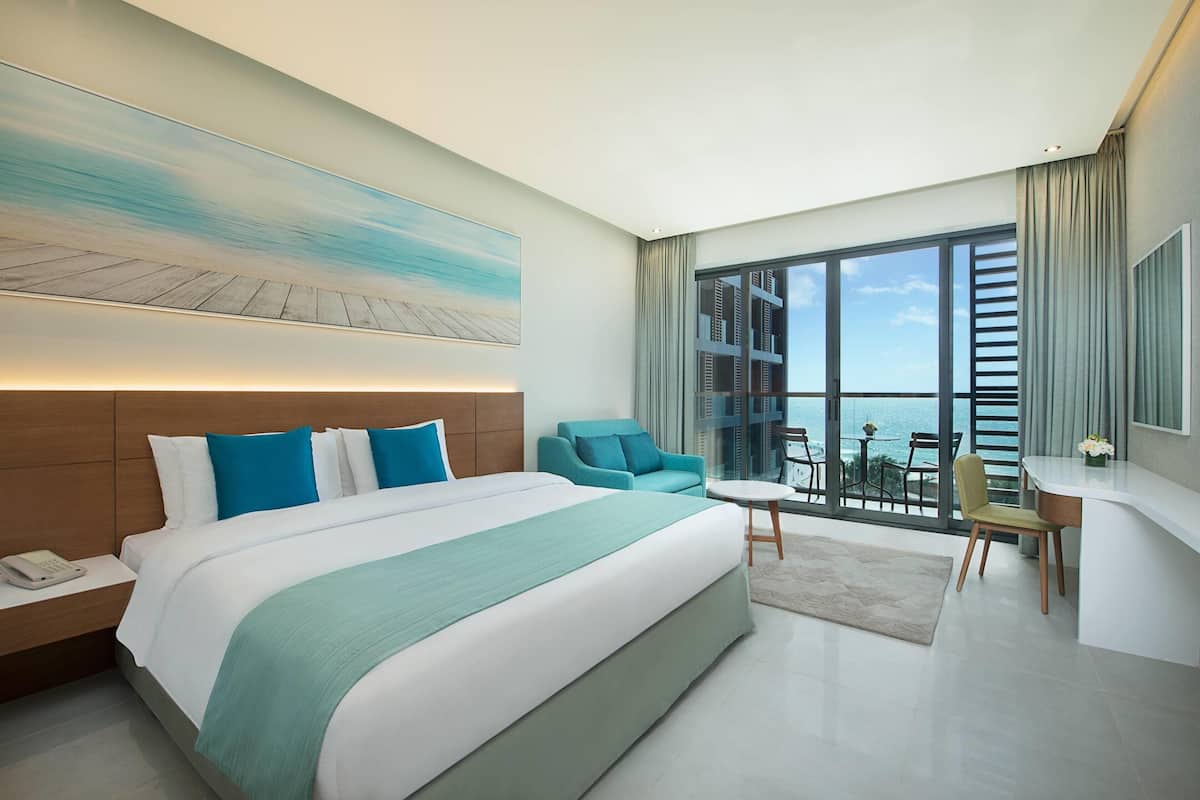 Een moderne hotelkamer met een groot bed, een bureau en uitzicht op de oceaan door grote ramen.