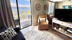 A living room with a large TV, a sofa, a chair, and a view of the beach through a sliding door.
