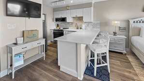 Une cuisine compacte avec des meubles blancs, un four à micro-ondes et une cuisinière. On y trouve un petit îlot avec une chaise, un téléviseur à écran plat fixé au mur, et un lit avec une tête de lit blanche dans la pièce adjacente.