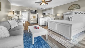 Un salon moderne avec un canapé gris, une table basse blanche et un tapis bleu. On aperçoit, au fond, un lit blanc avec une tête de lit décorative, un ventilateur de plafond et un coin cuisine.