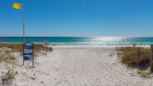 Une plage de sable avec un drapeau, un panneau indiquant les consignes de sécurité concernant le drapeau de plage, et un ciel bleu dégagé.