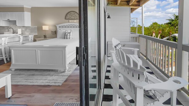 Une chambre avec un grand lit, un bureau et une chaise, une salle de bain avec des toilettes et un lavabo, et un balcon avec des chaises blanches et une vue sur la verdure.