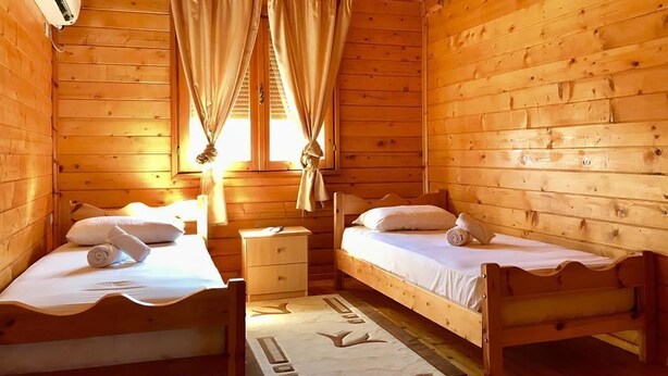 A wooden cabin room with two beds, a nightstand, and a window with curtains.