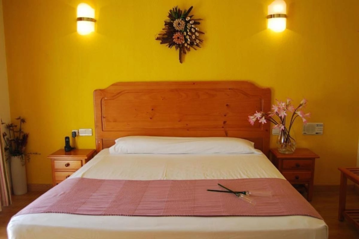 A hotel room with a wooden headboard, a bed with white linens, a pink throw, a bedside table with a vase of flowers, and wall-mounted lights.