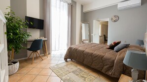 Un dormitorio con cama, escritorio, silla, televisión, espejo y planta.