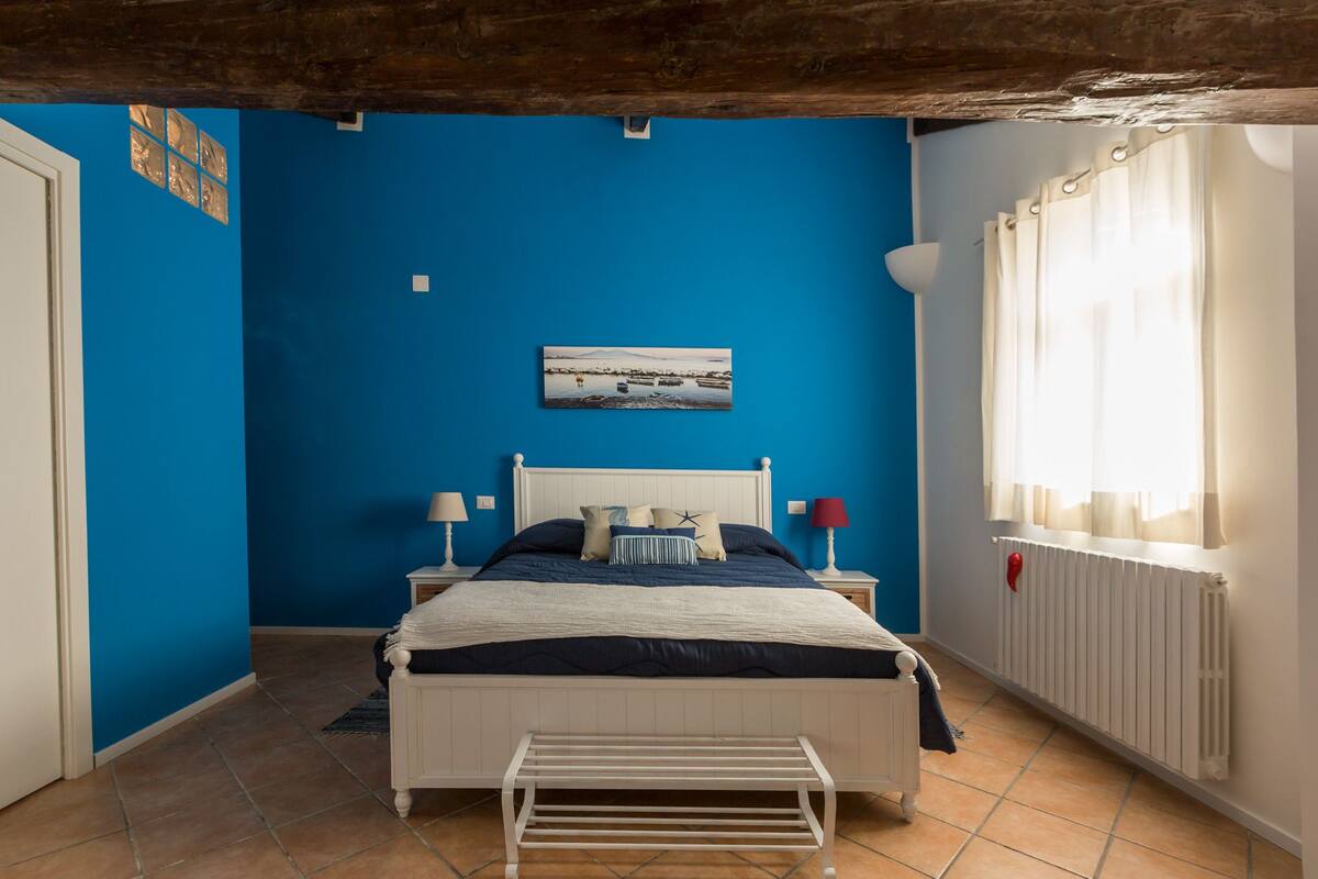Ein Schlafzimmer mit blauer Wand, einem Bett mit weißem Kopfteil, einem Nachttisch mit Lampe und einem Fenster mit Vorhängen.