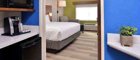 Habitación de hotel con cama, cafetera, microondas, lámpara, ventana y una planta pequeña.