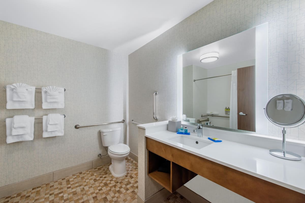 Une salle de bain moderne avec un lavabo blanc, un miroir et des toilettes. Des serviettes blanches sont suspendues sur un porte-serviettes, et le sol est recouvert d’un motif décoratif.