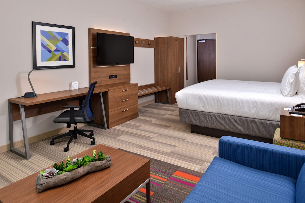 A hotel room with a bed, a desk with a chair, a television, a wardrobe, and a small seating area.