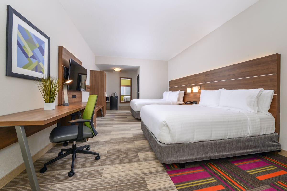 A hotel room with a desk, two beds, a chair, a TV, and a colorful rug.