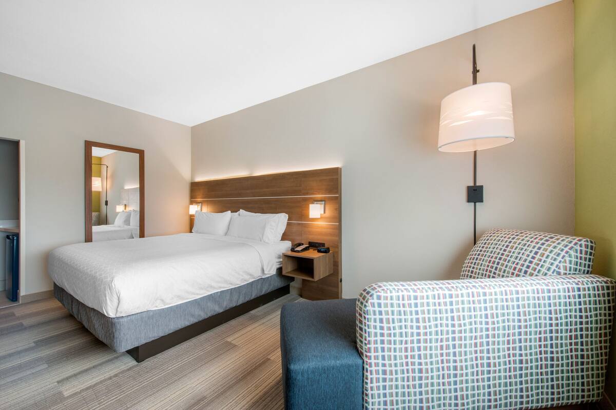 A hotel room with a bed, bedside table, lamp, and a chair with a patterned cover.