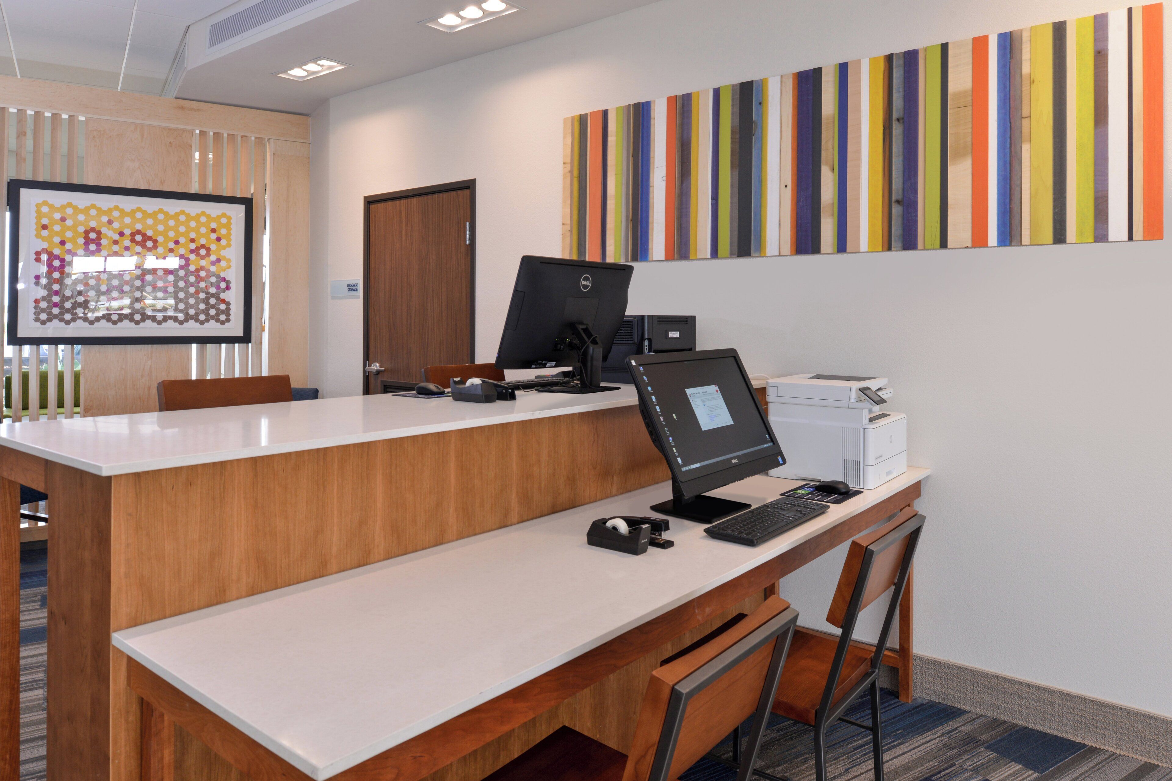 A modern office space with a long wooden desk, a computer, and a printer. There is a colorful artwork on the wall and a framed picture on the left.