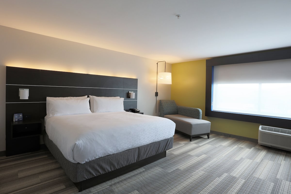A modern hotel room with a large bed, a bedside table, a chair, a window, and a wall-mounted air conditioner.