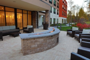 Un área de estar al aire libre con una chimenea de piedra, muebles de mimbre y un edificio con grandes ventanales al fondo.