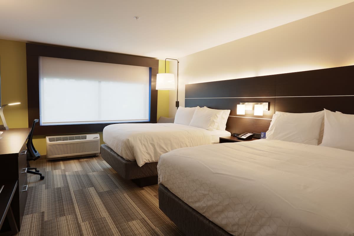 A hotel room with two beds, a desk, a chair, a large window with blinds, and an air conditioning unit.