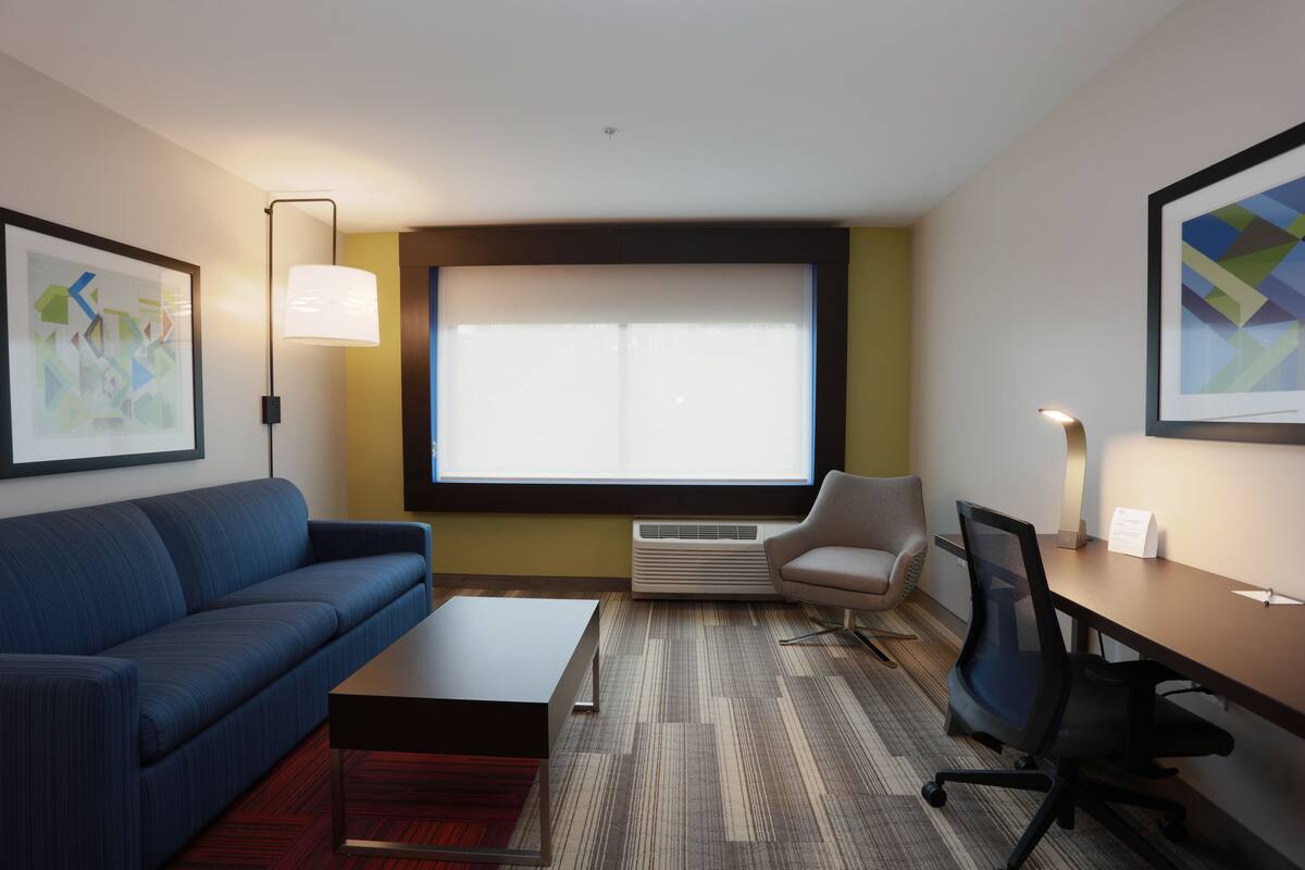 A modern hotel room with a blue sofa, a small table, a desk with a chair, a large window with blinds, and a framed abstract painting on the wall.