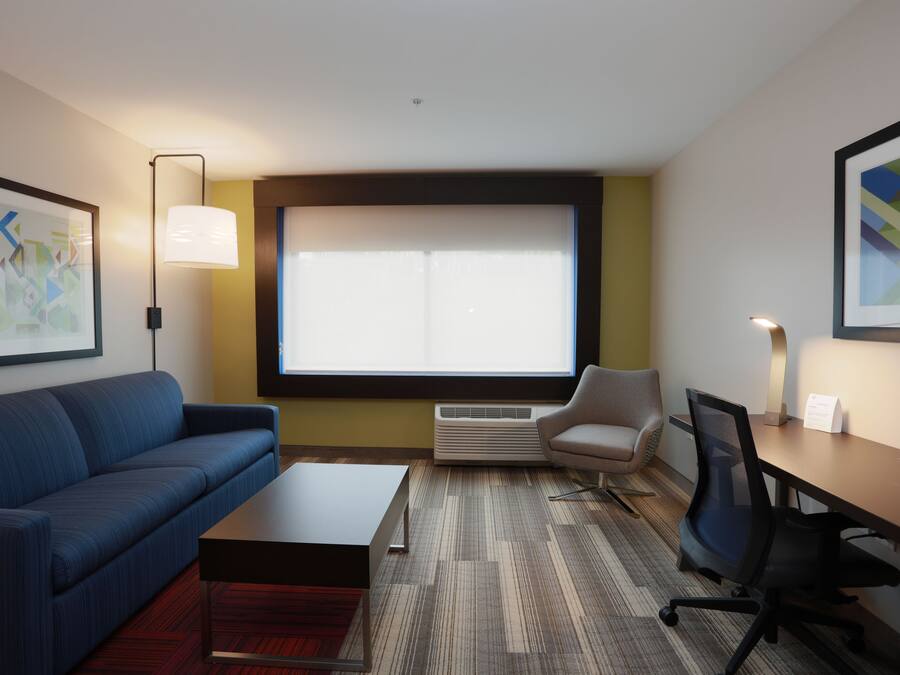 Een moderne hotelkamer met een blauwe bank, een klein tafeltje, een bureau met stoel, een groot raam met jaloezieën en een lijst met abstract schilderij aan de muur.