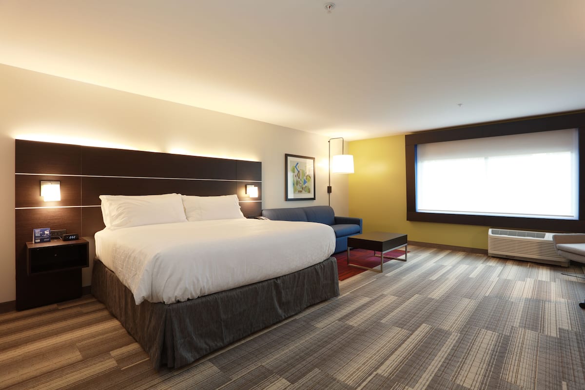 A modern hotel room with a large bed, a sofa, a desk, and a large window.