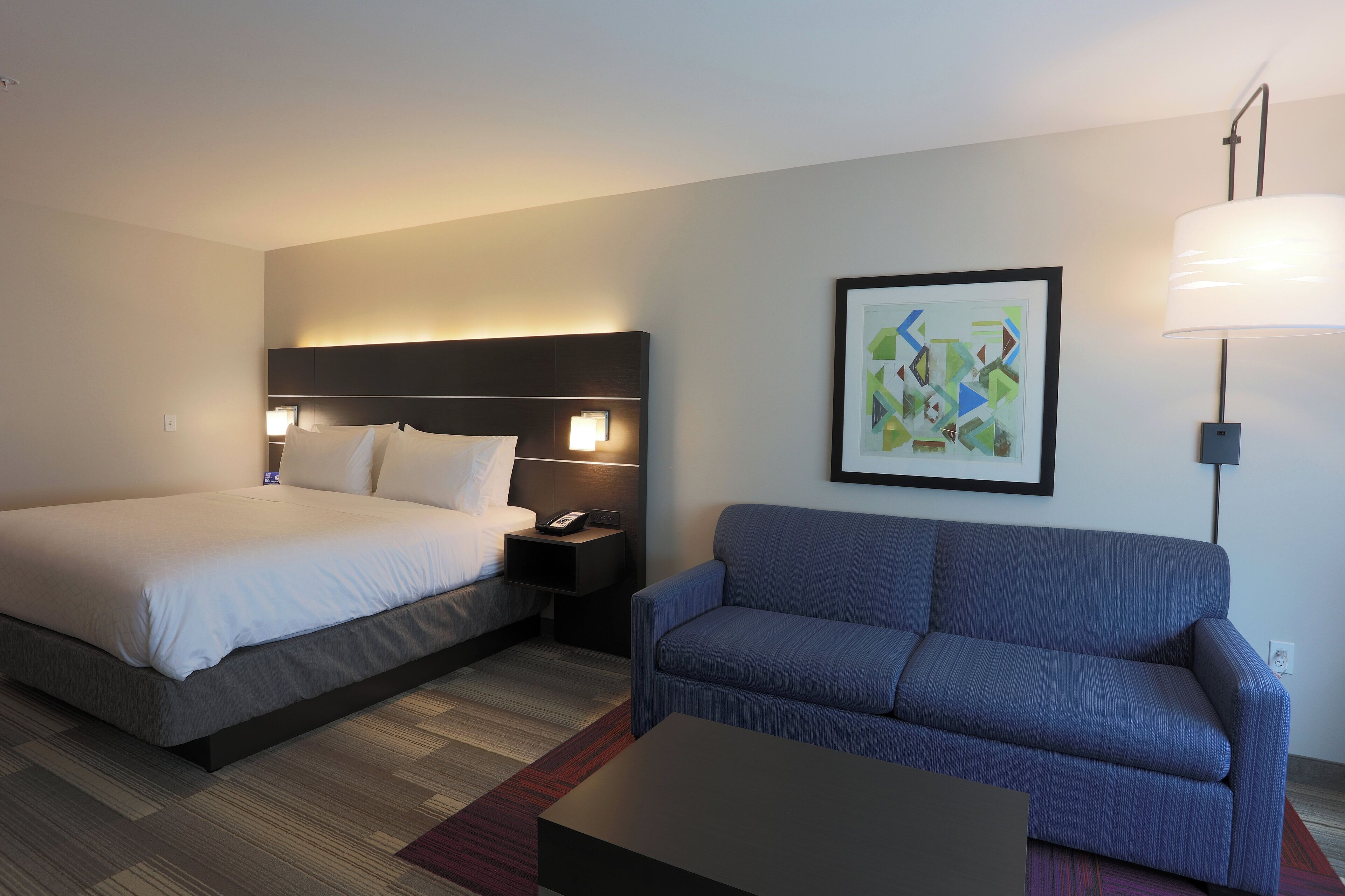 A hotel room with a bed, a sofa, a nightstand with a lamp, and a framed picture on the wall.