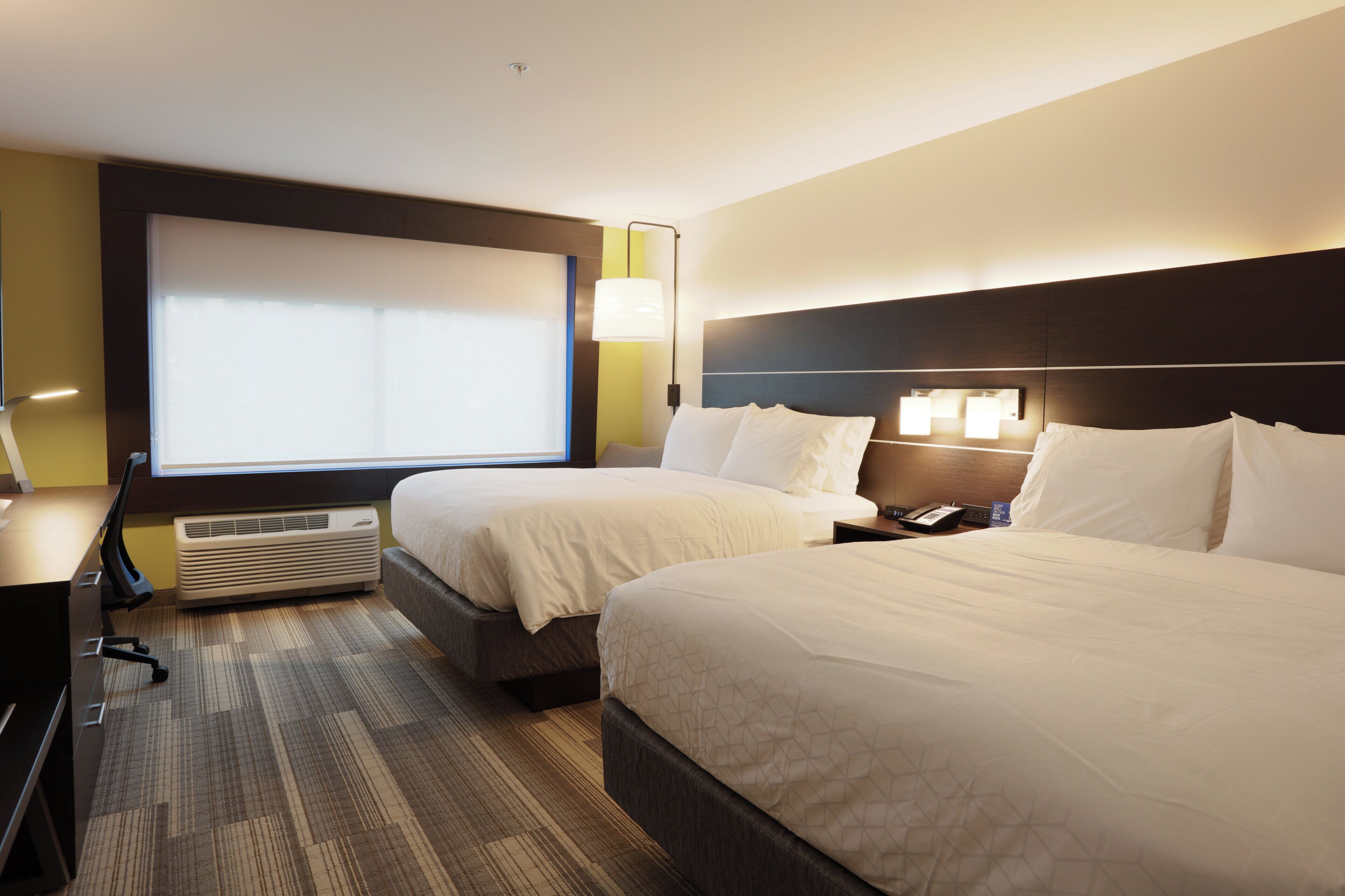 A hotel room with two beds, a desk, a chair, a large window, and a wall-mounted air conditioner.
