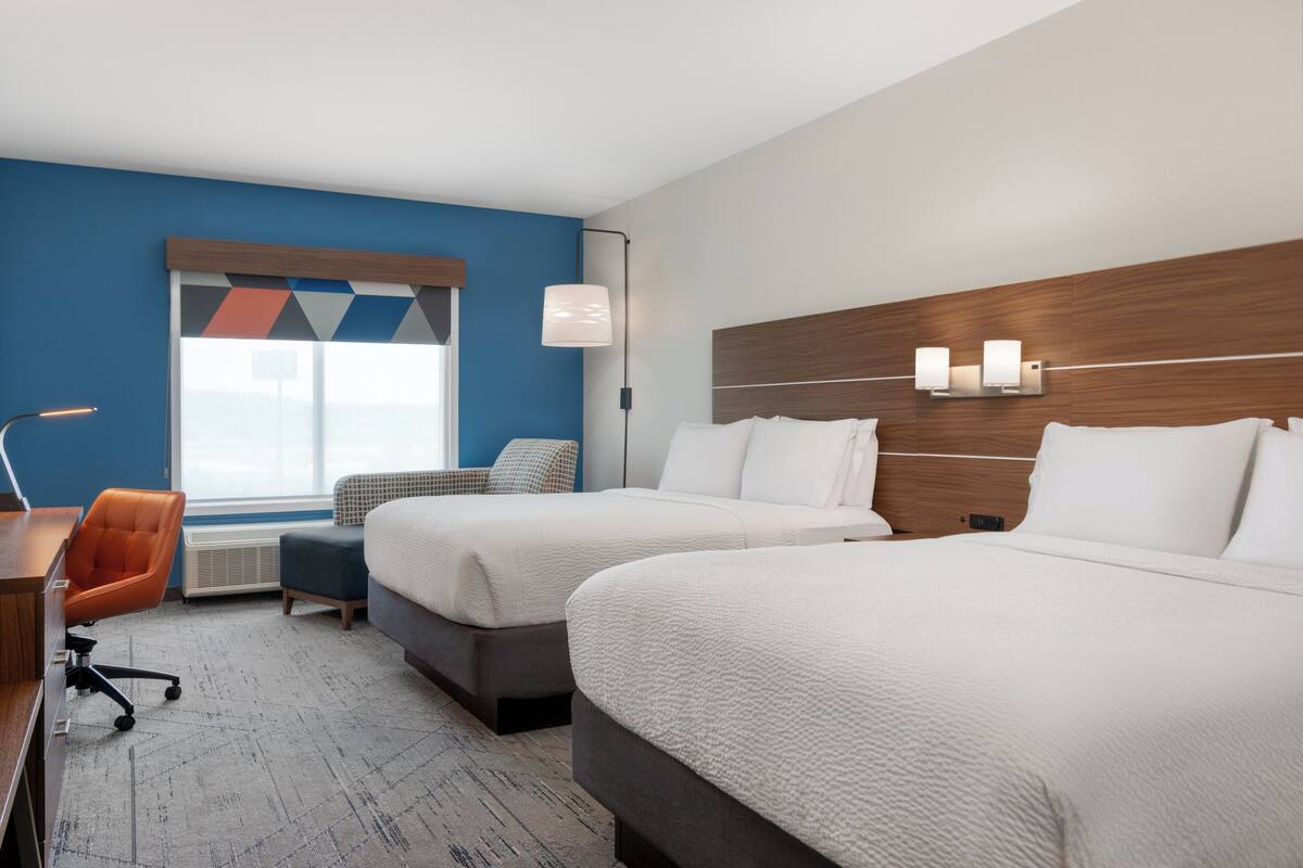 A hotel room with two beds, a desk, an orange chair, a window with a view, and a wall-mounted lamp.