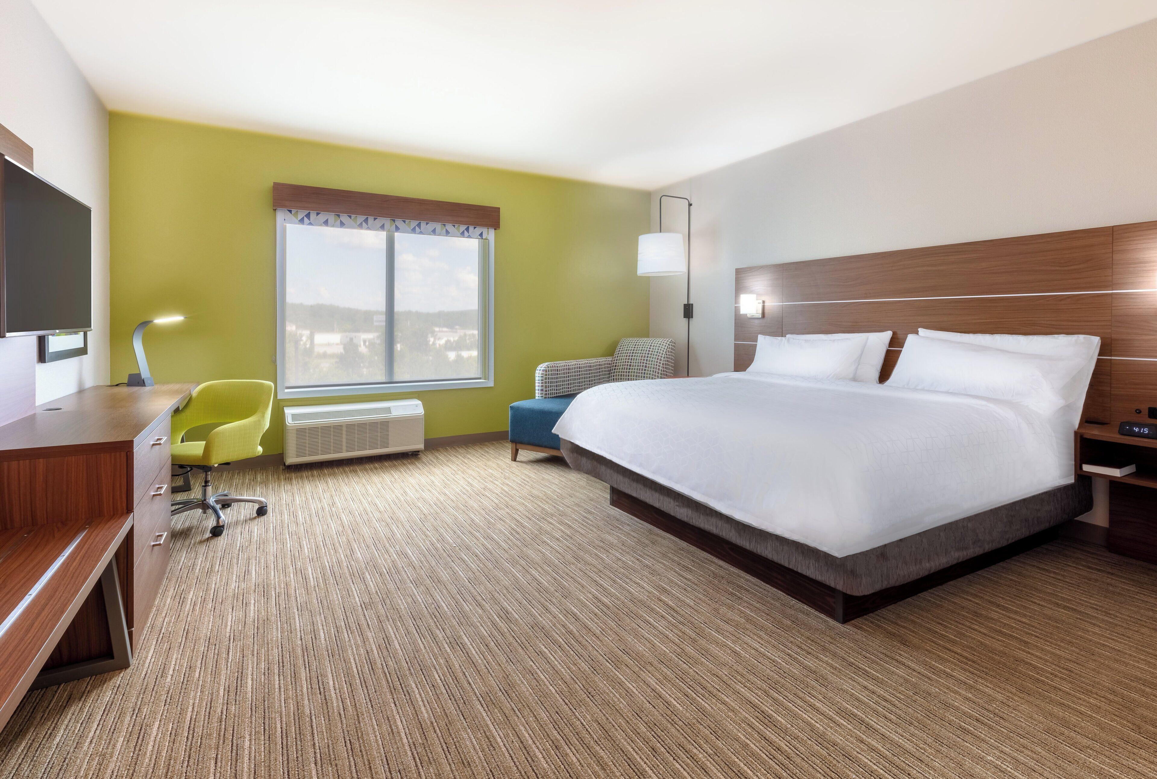 A hotel room with a bed, a desk, a chair, a TV, a window, and an air conditioning unit.