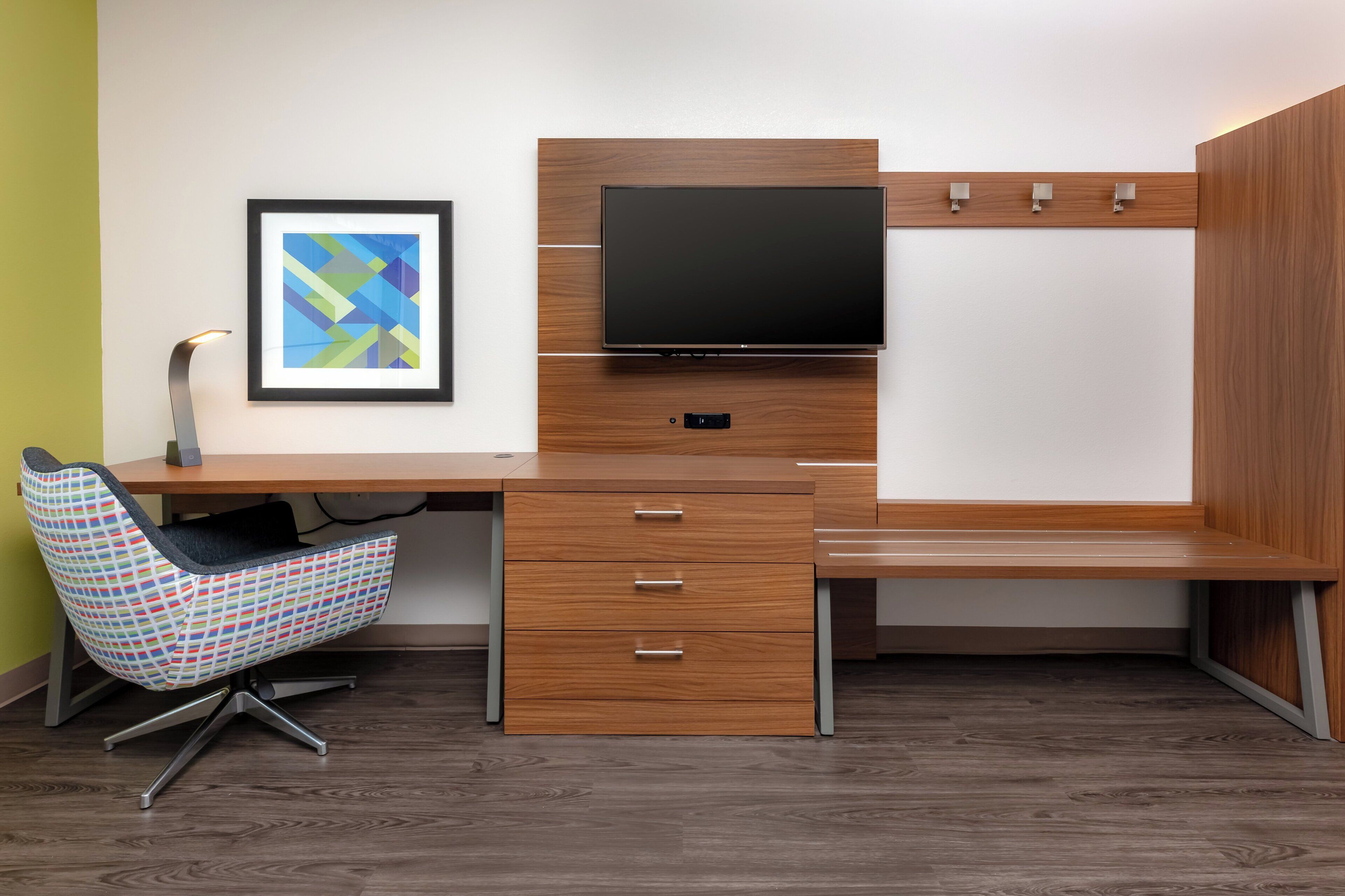 A modern office desk with a flat-screen monitor, a chair with a colorful pattern, and a framed abstract artwork on the wall.