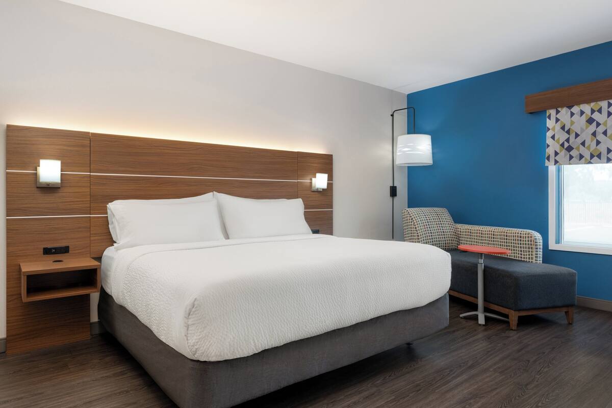A modern hotel room with a large bed, bedside tables, a chair, and a window with patterned curtains.