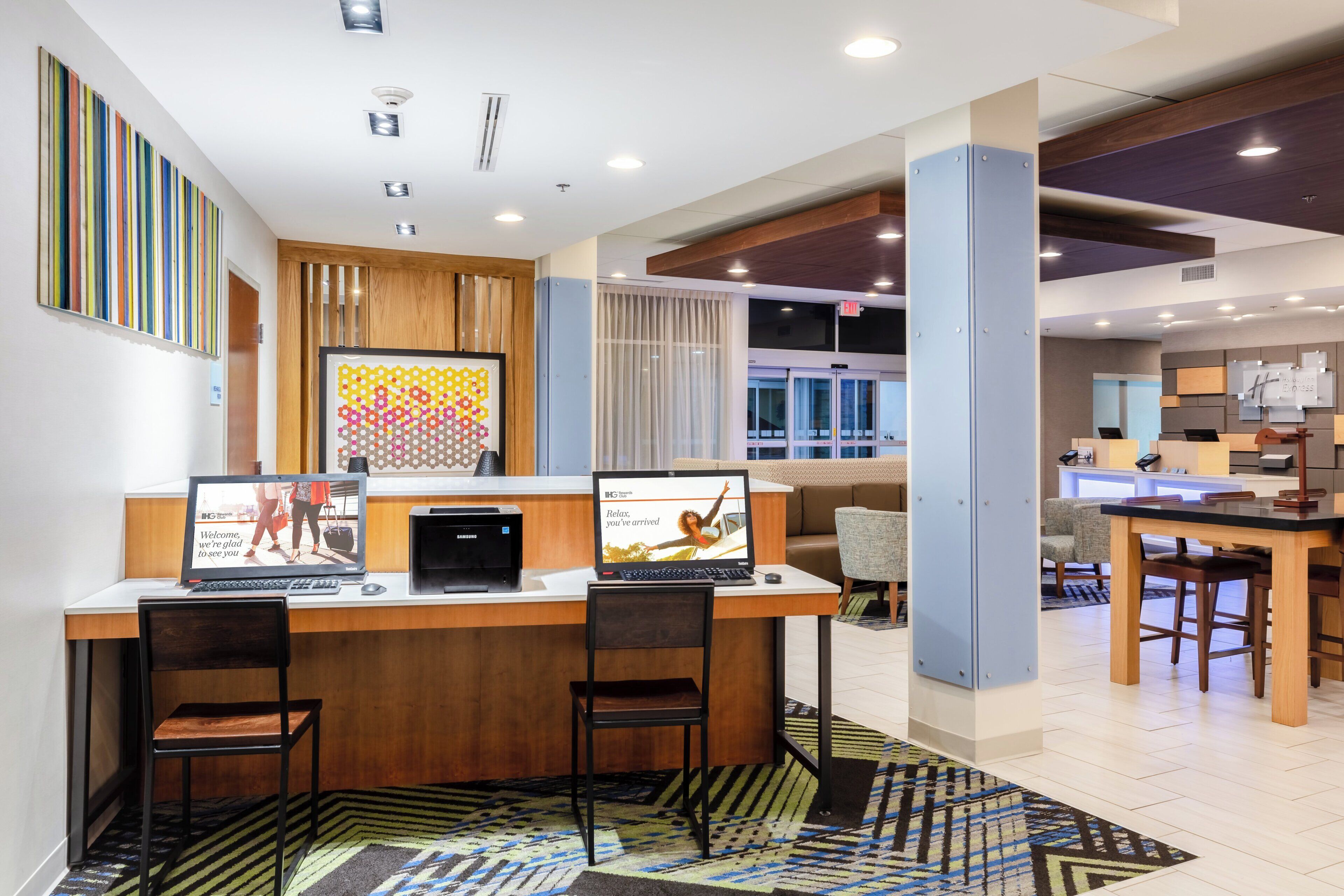 A hotel reception area with a wooden reception desk, two black chairs, a Samsung device, and a digital screen displaying a welcome message.
